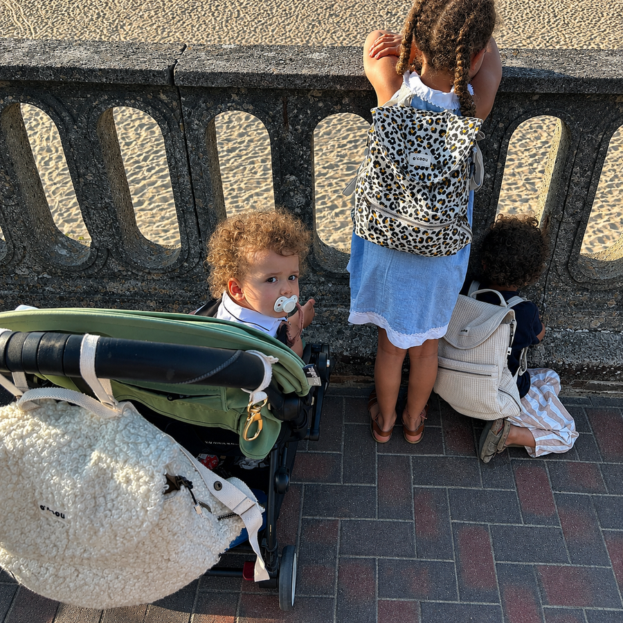 Deux enfants portent leur sac O'CAOU devant un muret en bord de plage, un bébé les regarde depuis sa poussette
