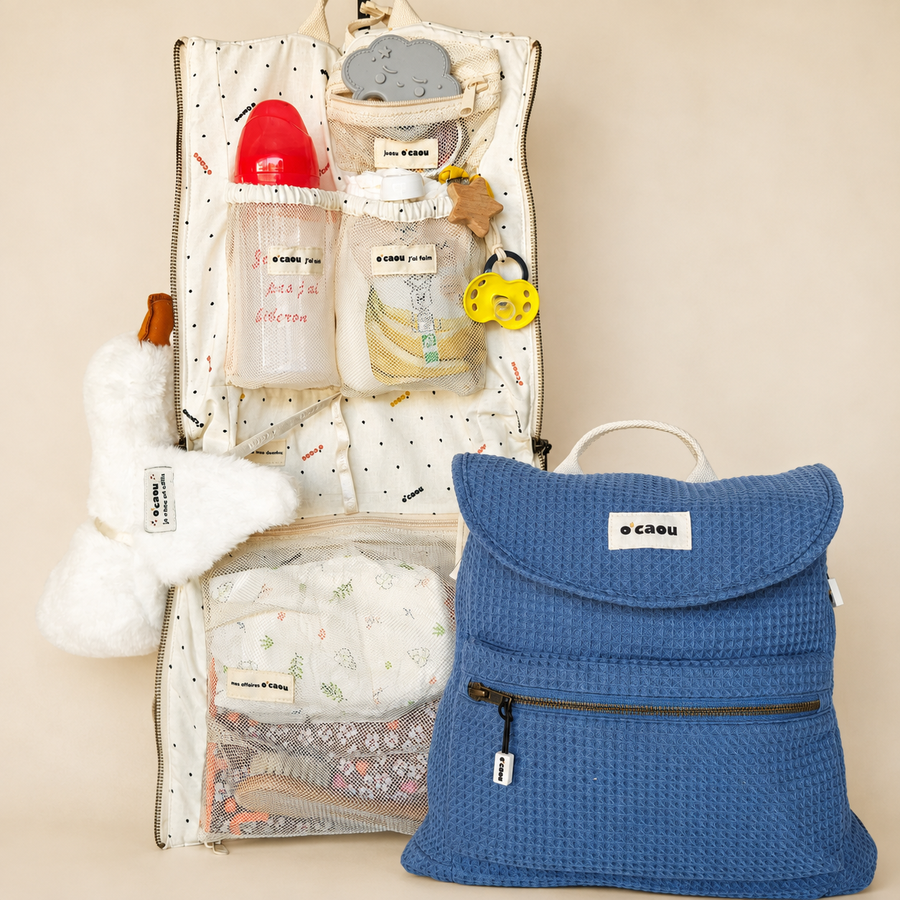 Sac O'CAOU bleu fermé, à côté de son organiseur déplié : biberon, tétine, doudou, couches et lange rangés dans les poches en filet